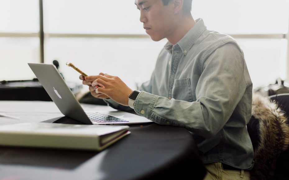 man looking down at phone while laptop is open