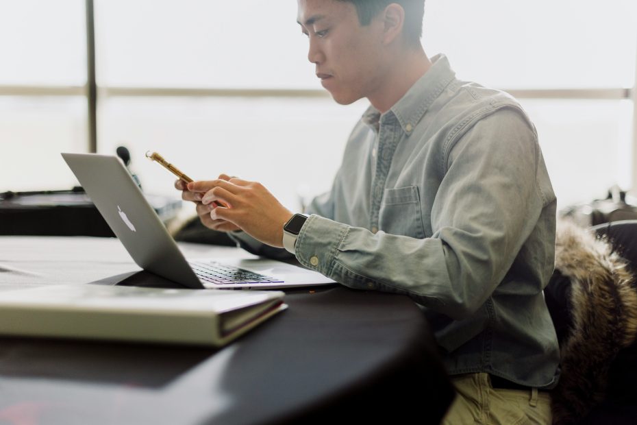 man looking down at phone while laptop is open